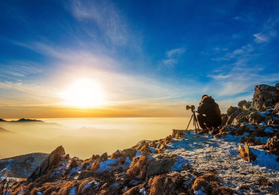 Professional photographer takes photos with camera on tripod on rocky peak at sunset.